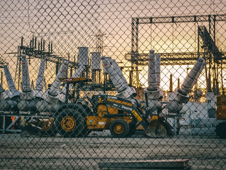An industrial power plant with heavy machinery and equipment behind a chain link fence during sunset, showcasing energy infrastructure.